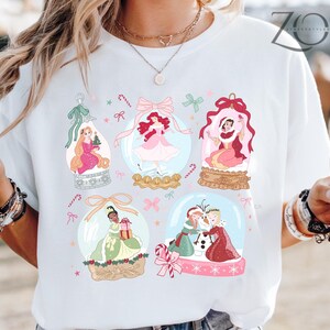 May include: White t-shirt featuring colorful illustrations of princesses inside snow globes. Each globe has a different princess, with candy canes and bows. The shirt has the text "ZOE" in the upper right corner.