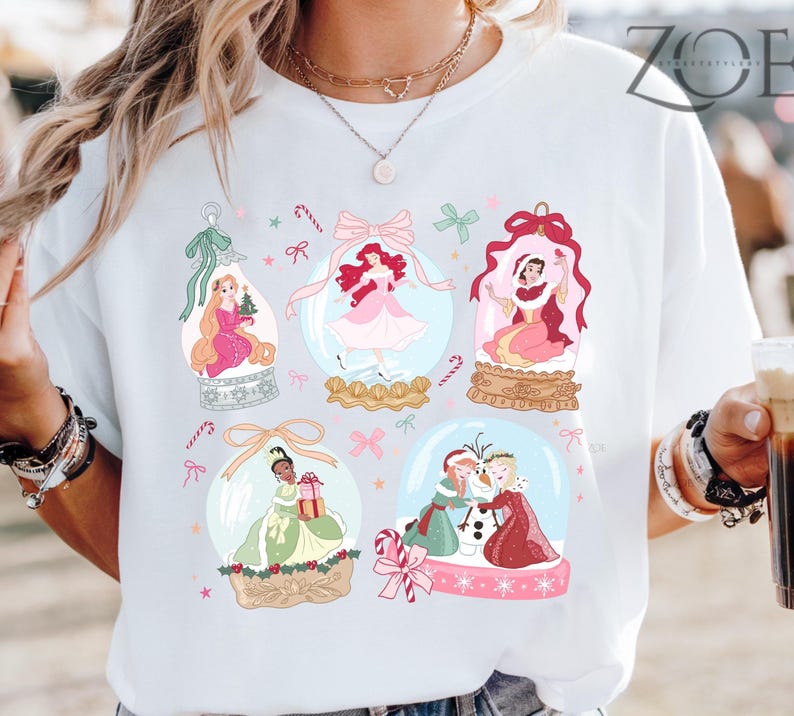 May include: White t-shirt featuring colorful illustrations of princesses inside snow globes. Each globe has a different princess, with candy canes and bows. The shirt has the text "ZOE" in the upper right corner.