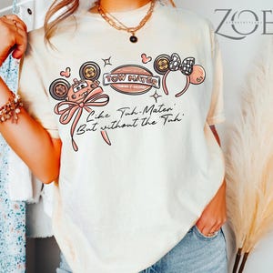 Może przedstawiać: Kremowa koszulka z grafiką Złomka z filmu Auta, z r&oacute;żową kokardą i napisem "Like Tuh-Mater But without the Tuh". Projekt zawiera uszy Myszki Miki i szachownicową kokardę.