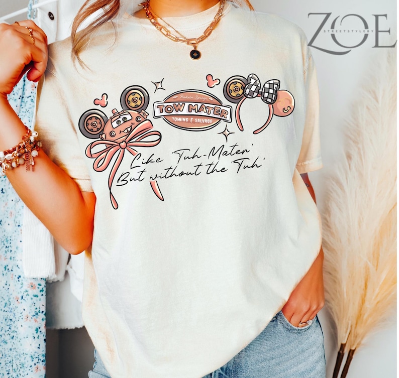 Może przedstawiać: Kremowa koszulka z grafiką Złomka z filmu Auta, z r&oacute;żową kokardą i napisem "Like Tuh-Mater But without the Tuh". Projekt zawiera uszy Myszki Miki i szachownicową kokardę.