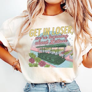 Puede incluir: Una camiseta color crema con un estampado gr&aacute;fico de un barco verde en un invernadero con hileras de lechuga. El texto dice "GET IN LOSER, we're learning about Lettuce". El dise&ntilde;o incluye peque&ntilde;as estrellas y detalles florales.
