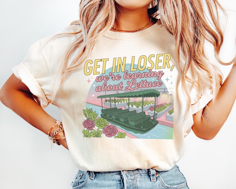 Puede incluir: Una camiseta color crema con un estampado gr&aacute;fico de un barco verde en un invernadero con hileras de lechuga. El texto dice "GET IN LOSER, we're learning about Lettuce". El dise&ntilde;o incluye peque&ntilde;as estrellas y detalles florales.