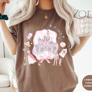 May include: Espresso-colored t-shirt featuring a whimsical castle design with pink, blue, and purple accents. The graphic includes a pink bow, a dress, and other fairytale-themed elements. The shirt is labeled "Comfort Colors Espresso".