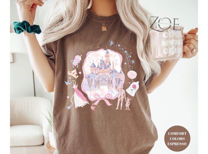 May include: Espresso-colored t-shirt featuring a whimsical castle design with pink, blue, and purple accents. The graphic includes a pink bow, a dress, and other fairytale-themed elements. The shirt is labeled "Comfort Colors Espresso".