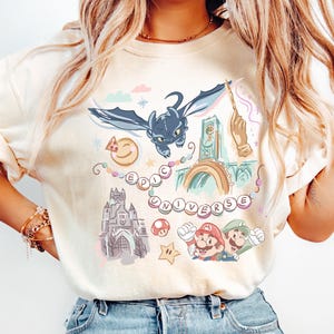 K&ouml;nnte beinhalten: Ein wei&szlig;es T-Shirt mit einem bunten Grafikdruck, der einen Drachen, ein Schloss, einen Uhrturm, einen Pilz, einen Stern und den Text "EPIC UNIVERSE" zeigt.