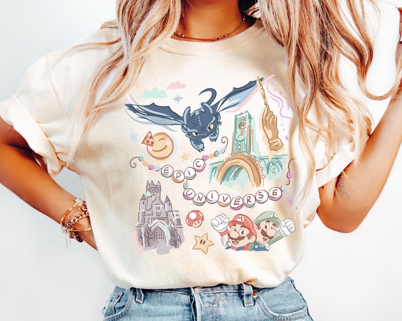 K&ouml;nnte beinhalten: Ein wei&szlig;es T-Shirt mit einem bunten Grafikdruck, der einen Drachen, ein Schloss, einen Uhrturm, einen Pilz, einen Stern und den Text "EPIC UNIVERSE" zeigt.