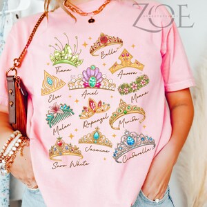 K&ouml;nnte beinhalten: Rosafarbenes T-Shirt mit bunten Illustrationen verschiedener Prinzessinnen-Diademe, die jeweils mit dem Namen einer Prinzessin beschriftet sind, darunter Tiana, Elsa, Arielle, Belle, Aurora, Moana, Mulan, Rapunzel, Merida, Schneewittchen, Jasmin und Cinderella.
