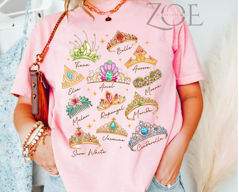 K&ouml;nnte beinhalten: Rosafarbenes T-Shirt mit bunten Illustrationen verschiedener Prinzessinnen-Diademe, die jeweils mit dem Namen einer Prinzessin beschriftet sind, darunter Tiana, Elsa, Arielle, Belle, Aurora, Moana, Mulan, Rapunzel, Merida, Schneewittchen, Jasmin und Cinderella.
