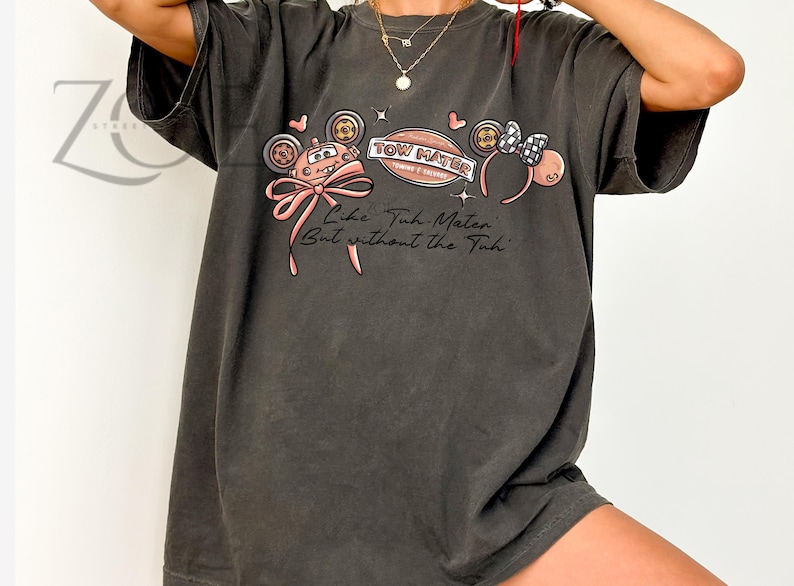 Może przedstawiać: Ciemnoszara koszulka z nadrukiem przedstawiającym samoch&oacute;d, kokardę i opaskę na głowę. Tekst na koszulce brzmi "Tow Mater" i "Like Tow Mater But without the Tuh". Projekt zawiera r&oacute;żowe i czarne akcenty.