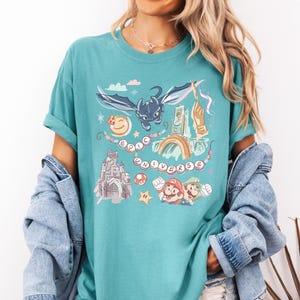K&ouml;nnte beinhalten: Ein t&uuml;rkisfarbenes T-Shirt mit einem grafischen Aufdruck eines schwarzen Drachen, eines Schlosses, eines Regenbogens und Figuren aus einem beliebten Videospiel. Der Text "EPIC UNIVERSE" ist auf dem Shirt aufgedruckt.