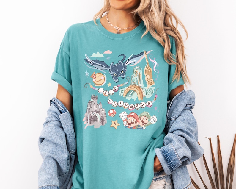 K&ouml;nnte beinhalten: Ein t&uuml;rkisfarbenes T-Shirt mit einem grafischen Aufdruck eines schwarzen Drachen, eines Schlosses, eines Regenbogens und Figuren aus einem beliebten Videospiel. Der Text "EPIC UNIVERSE" ist auf dem Shirt aufgedruckt.