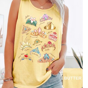 May include: Yellow tank top featuring a collection of illustrated princess tiaras. Each tiara is unique in design and color, with names like Ariel, Belle, and Cinderella written below. The shirt has a round neckline.
