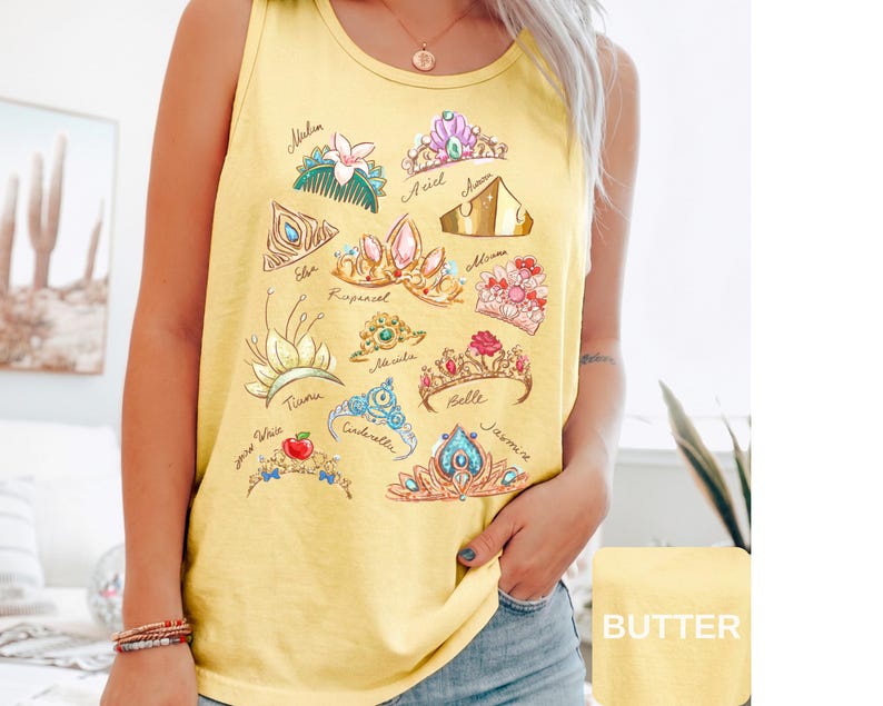 May include: Yellow tank top featuring a collection of illustrated princess tiaras. Each tiara is unique in design and color, with names like Ariel, Belle, and Cinderella written below. The shirt has a round neckline.