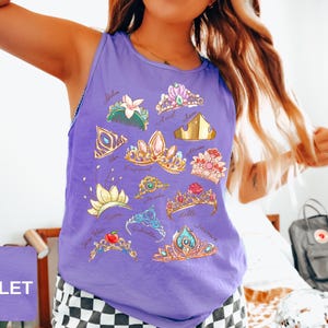 May include: A violet tank top featuring colorful illustrations of various tiaras and crowns. The designs include gold, pink, blue, and green accents, with names like Ariel, Jasmine, and Cinderella. The word "Violet" is in a violet square in the bottom left corner.