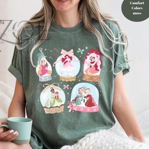 May include: Green t-shirt featuring illustrations of princesses inside snow globes. The design includes pink, red, and gold accents, with the text "Comfort Colors moss" in a circle on the upper right.