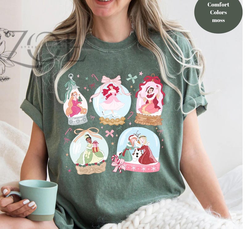 May include: Green t-shirt featuring illustrations of princesses inside snow globes. The design includes pink, red, and gold accents, with the text "Comfort Colors moss" in a circle on the upper right.