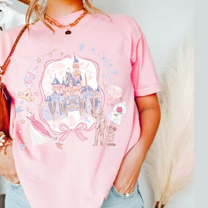 May include: Pink t-shirt featuring a whimsical illustration of a fairytale castle, a bow, and other themed graphics. The design includes a castle, a rose in a glass dome, and a couple. The shirt is a soft pink color.