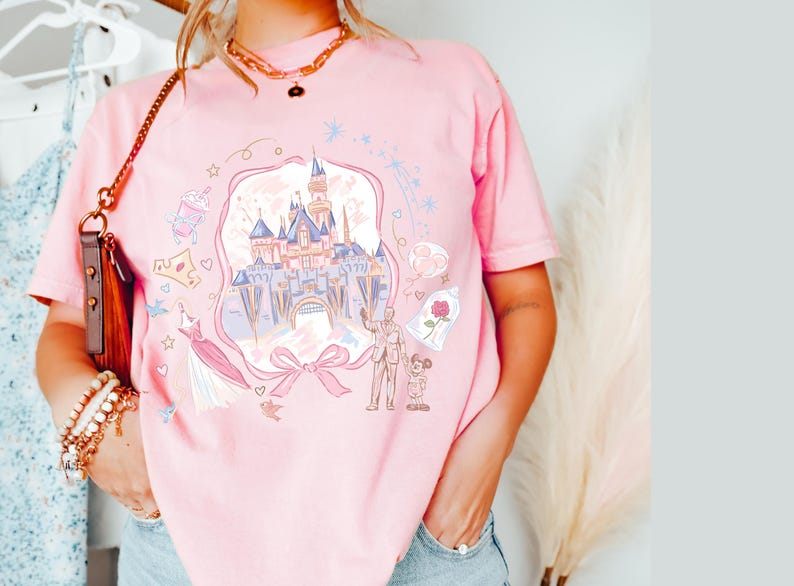May include: Pink t-shirt featuring a whimsical illustration of a fairytale castle, a bow, and other themed graphics. The design includes a castle, a rose in a glass dome, and a couple. The shirt is a soft pink color.