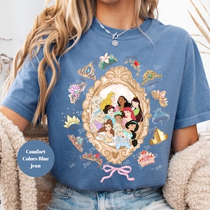 May include: Blue jean-colored t-shirt featuring a gold-framed portrait of animated princesses. The portrait is surrounded by tiaras and crowns. A pink bow is at the bottom of the frame. The text "Comfort Colors Blue jean" is on the shirt.