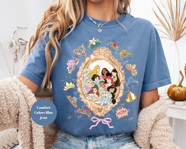 May include: Blue jean-colored t-shirt featuring a gold-framed portrait of animated princesses. The portrait is surrounded by tiaras and crowns. A pink bow is at the bottom of the frame. The text "Comfort Colors Blue jean" is on the shirt.