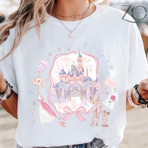 May include: White t-shirt featuring a pastel illustration of a fairytale castle, surrounded by whimsical elements like a crown, dress, and characters. The design is framed by a pink ribbon and bow.