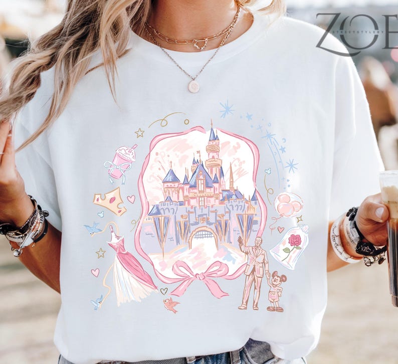 May include: White t-shirt featuring a pastel illustration of a fairytale castle, surrounded by whimsical elements like a crown, dress, and characters. The design is framed by a pink ribbon and bow.
