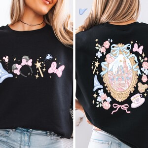 May include: Black sweatshirt featuring a whimsical design with a light blue wizard hat, pink bow, and a wand on the front. The back showcases a castle in a gold frame surrounded by flowers, bows, and a teacup.