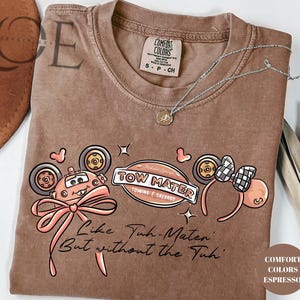 Może przedstawiać: Brązowa koszulka w kolorze espresso z grafiką Złomka z filmu Auta, kokardą i napisem "Like Tuh-Mater But without the Tuh". Koszulka posiada r&oacute;wnież parę uszu Myszki Miki.