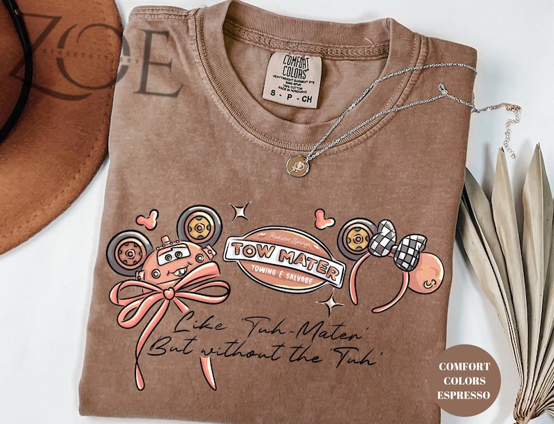 Może przedstawiać: Brązowa koszulka w kolorze espresso z grafiką Złomka z filmu Auta, kokardą i napisem "Like Tuh-Mater But without the Tuh". Koszulka posiada r&oacute;wnież parę uszu Myszki Miki.