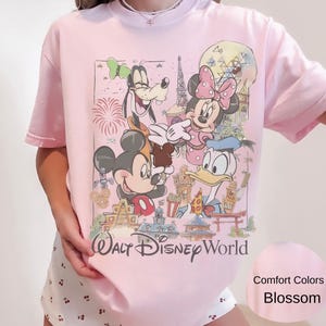 May include: A light pink t-shirt featuring a graphic print of Mickey Mouse, Minnie Mouse, Goofy, and Donald Duck in front of Walt Disney World landmarks. The text Walt Disney World is printed in black below the characters. The shirt is short-sleeved.