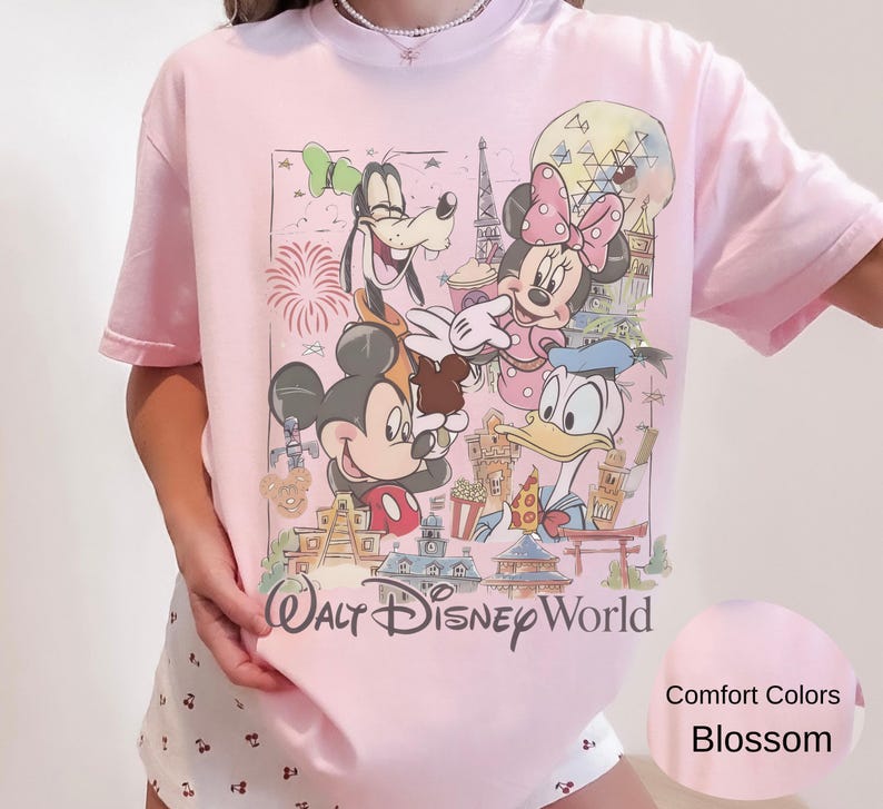 May include: A light pink t-shirt featuring a graphic print of Mickey Mouse, Minnie Mouse, Goofy, and Donald Duck in front of Walt Disney World landmarks. The text Walt Disney World is printed in black below the characters. The shirt is short-sleeved.
