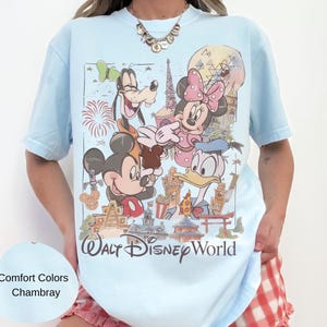 May include: A light blue t-shirt featuring a graphic print of Mickey Mouse, Minnie Mouse, Goofy, and Donald Duck in front of Walt Disney World landmarks. The text Walt Disney World is printed at the bottom of the graphic in a dark brown script font.