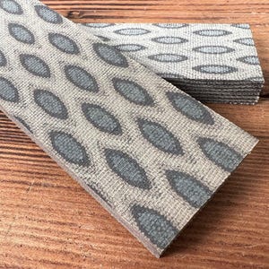 May include: A rectangular strip of textured material featuring a repeating pattern of dark gray, almond-shaped ovals set against a light gray, woven-look background. The strip has a visible layered edge and a flat, firm surface structure.