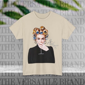 Puede incluir: Una camiseta beige con un estampado gráfico de Adele con rulos azules en el pelo y un jersey de cuello alto negro. Ella está sosteniendo una copa de vino en su boca y su firma está escrita en tinta negra debajo de ella. El texto "The Versatile Brand" se repite en el fondo.