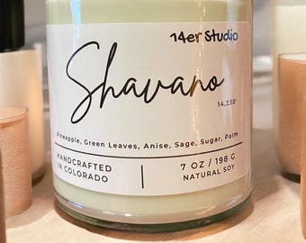 Op de afbeelding: Een witte kaars pot met een groen label. Het label zegt "14er Studio", "Shavano", "14,230", "Pineapple, Green Leaves, Anise, Sage, Sugar, Palm", "HANDCRAFTED IN COLORADO", en "7 OZ/198 G NATURAL SOY".