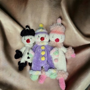 May include: Three crocheted stuffed toys in the shape of clowns. The clown on the left is black and white, the middle clown is purple and white, and the clown on the right is pink and white.