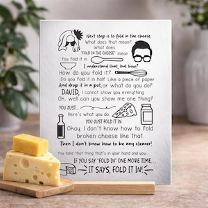 May include: A white rectangular sign with black text and illustrations about folding cheese, with two cheese wedges on a wooden board in front. The sign includes the text "FOLD IN THE CHEESE" and humorous instructions.