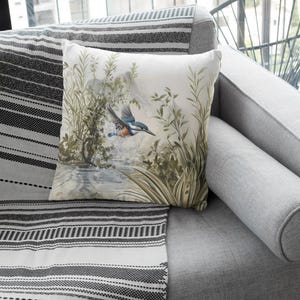 May include: A white throw pillow with a watercolor illustration of a blue kingfisher bird flying over a stream with green foliage.