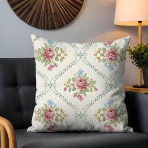 May include: Square throw pillow with a floral pattern. The pillow features a cream-colored background with a repeating pattern of pink roses, blue flowers, and green leaves. The design includes a trellis-like pattern with bows.