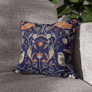 May include: Square decorative pillow with a navy blue background and a floral and bird pattern. The design features orange, beige, and white flowers, with two birds. The pillow is placed on a gray sofa.