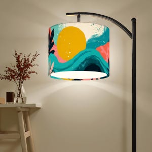May include: A modern floor lamp with a black metal frame and a cylindrical lampshade. The lampshade features an abstract design with a large yellow sun, teal waves, and pink and black floral elements.