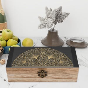 May include: A rectangular wooden box with a light brown wood grain finish and a dark gray lid featuring a gold-colored, semi-circular fan-shaped pattern with intricate line work and a central antique brass-toned metal latch on the front.