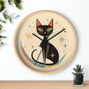 May include: A round wall clock with a mid-century modern design. The clock face features a black cat illustration with green eyes, set against a cream background. The clock has a wooden frame and black clock hands. The cat is sitting on a colorful crescent moon.