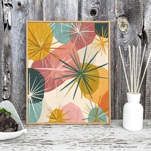 May include: Abstract art print with a mid-century modern design. The artwork features colorful shapes in shades of yellow, pink, teal, and green, with starburst-like elements. The print is framed in a light wood frame.