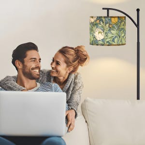 May include: A black floor lamp with a floral patterned lampshade. The lampshade features a design of white peonies, yellow flowers, and green leaves on a blue background. The lamp is positioned near a couple sitting on a white couch.