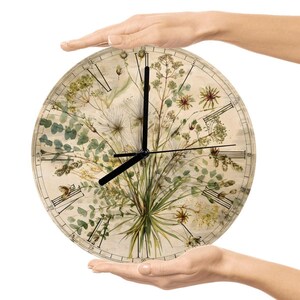 May include: Round wall clock featuring a botanical design with green and beige tones. The clock face displays Roman numerals and black clock hands. The clock is held by two hands against a white background.