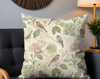May include: Square throw pillow with a vintage floral and bird pattern. The design features pink roses, green foliage, and various birds on a cream-colored background. The pillow is placed on a dark gray couch.