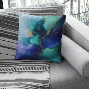 May include: A throw pillow featuring an abstract floral design in shades of blue, green and gold. The pillow is placed on a grey sofa with a black and white striped throw blanket.