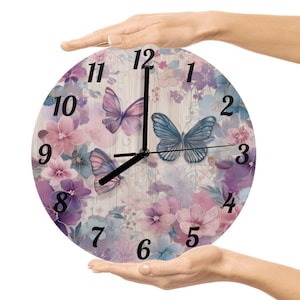 May include: Round wall clock featuring a floral and butterfly design in shades of purple, pink, and blue. The clock face has black numerals and hands. The clock is held by two hands.