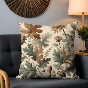 May include: Square cushion with a botanical design featuring oak leaves, chrysanthemums, and mushrooms in shades of brown, green, and beige. The cushion is placed on a dark grey sofa.